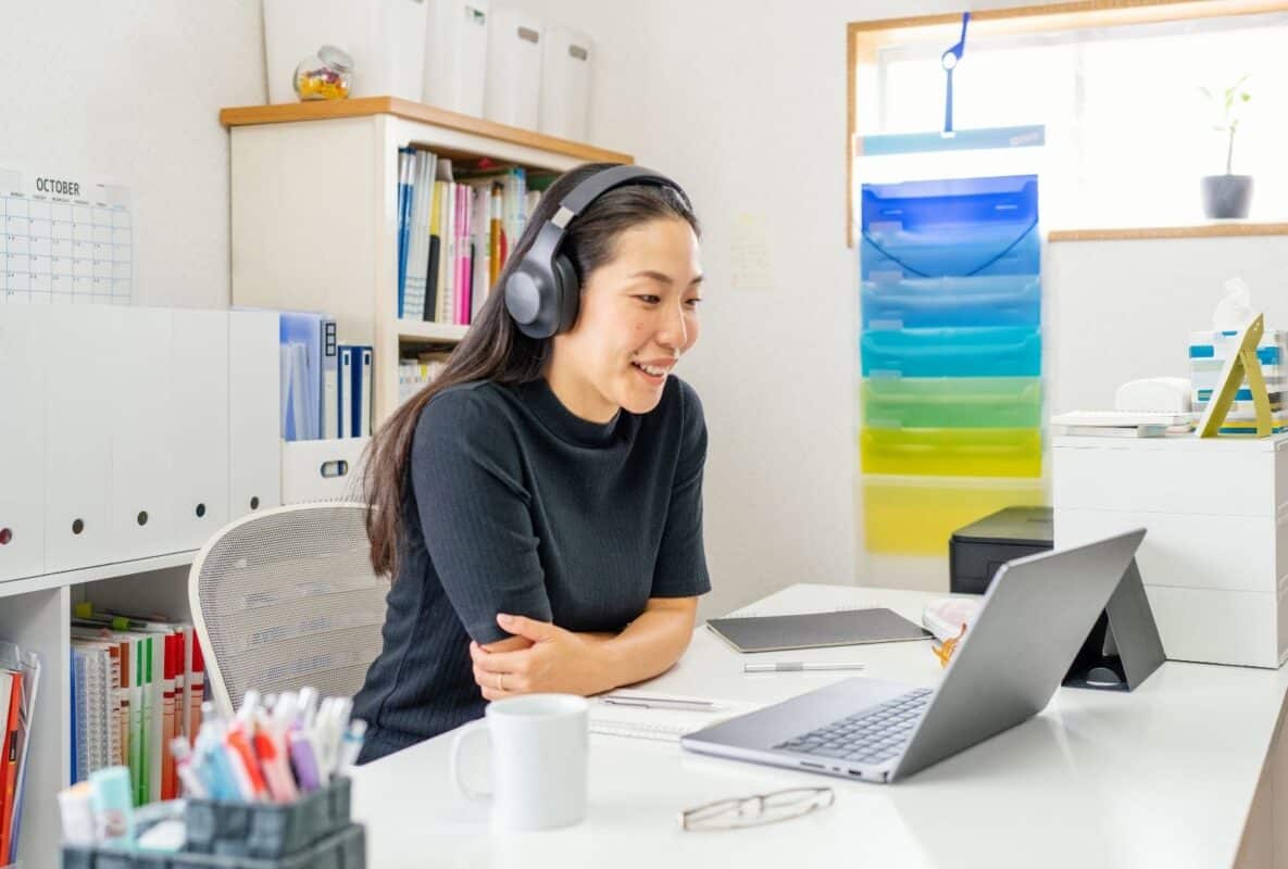 Smiling Asian student in cozy home office, wearing headphones for online Hebrew lesson with tutor Anat on laptop. Books, calendar, and tea enhance focused learning vibe.