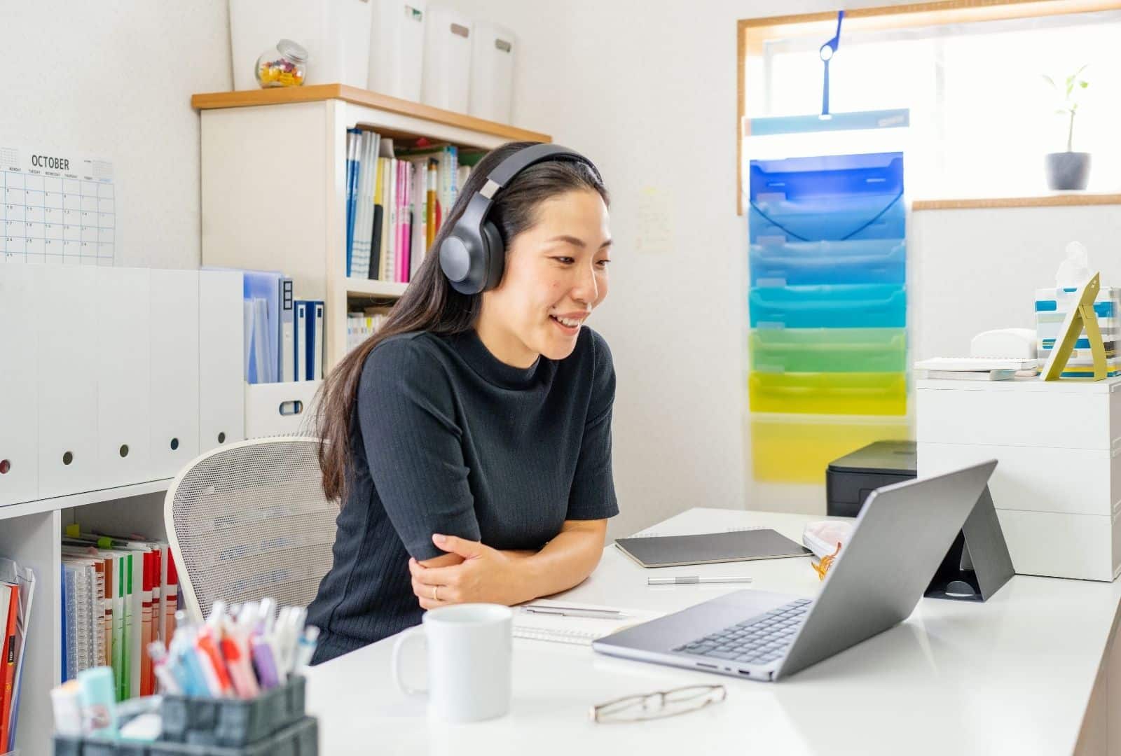 Smiling Asian student in cozy home office, wearing headphones for online Hebrew lesson with tutor Anat on laptop. Books, calendar, and tea enhance focused learning vibe.