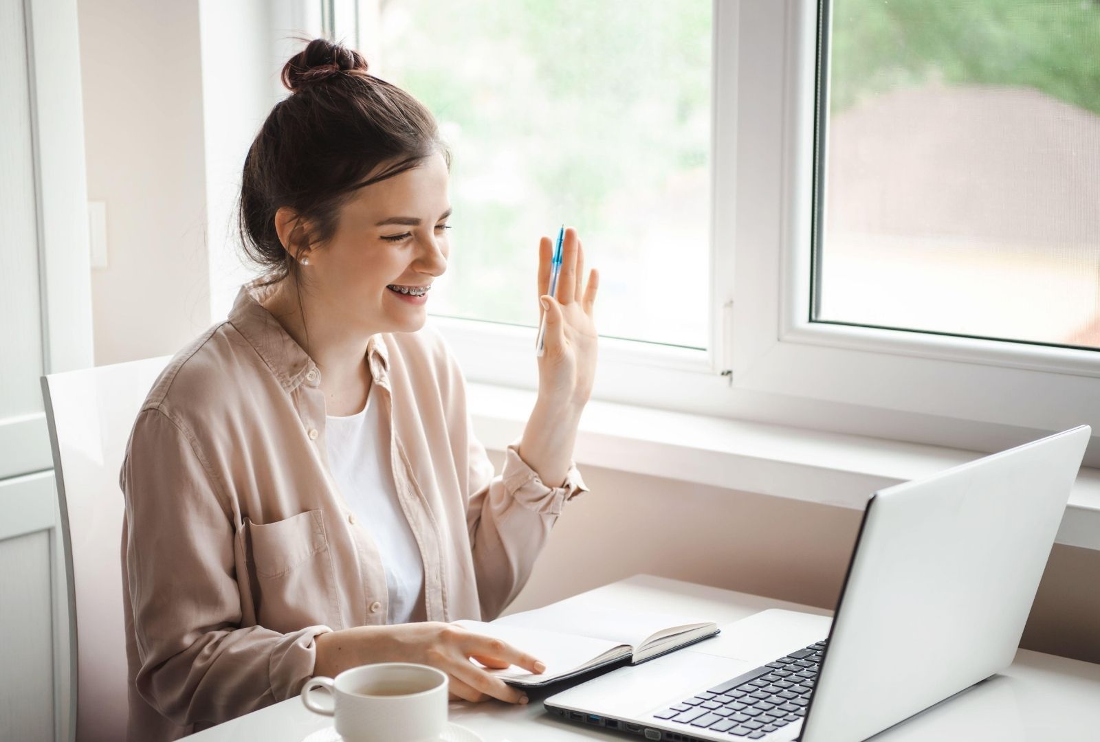 Happy student of online Hebrew teacher Anat Migdalor, smiling and waving with blue pen during engaging Zoom lesson—laptop, notebook, and coffee on desk by sunny window, embodying laughter in Hebrew learning.
