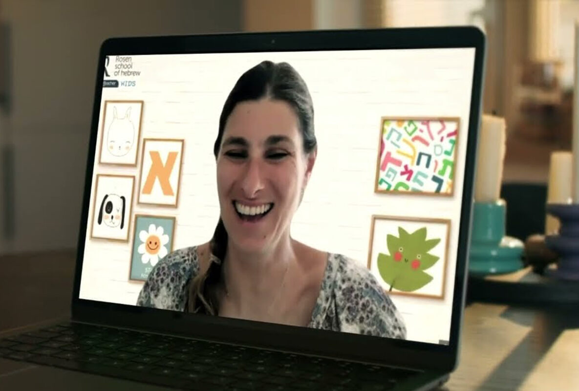 Hebrew Teacher Anat smiling in fun online Hebrew class at Rosen School, with colorful art frames of animals and letters.