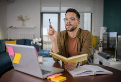 Young man studying English online at home desk with laptop, notebook, and pen—ideal for Anat Megidlor's virtual lessons.