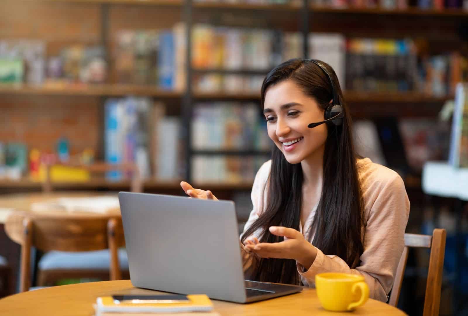 A smiling young woman with long dark hair, wearing wireless headphones and a light pink blouse, engages in an online video call from a cozy wooden desk in a sunlit library filled with bookshelves. She gestures animatedly with her hands while facing a silver laptop screen, with a yellow mug of tea and notebooks nearby, evoking enthusiasm for virtual language learning.