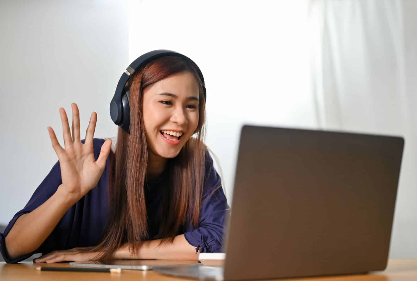 A cheerful young woman with long brown hair, wearing black wireless headphones and a navy blue blouse, waves enthusiastically with an open hand while smiling at her laptop screen during an online video call for an Intensive Hebrew Course Online. She's seated at a wooden desk in a bright library surrounded by wooden bookshelves filled with colorful books and green potted plants, capturing the joy of virtual language lessons.