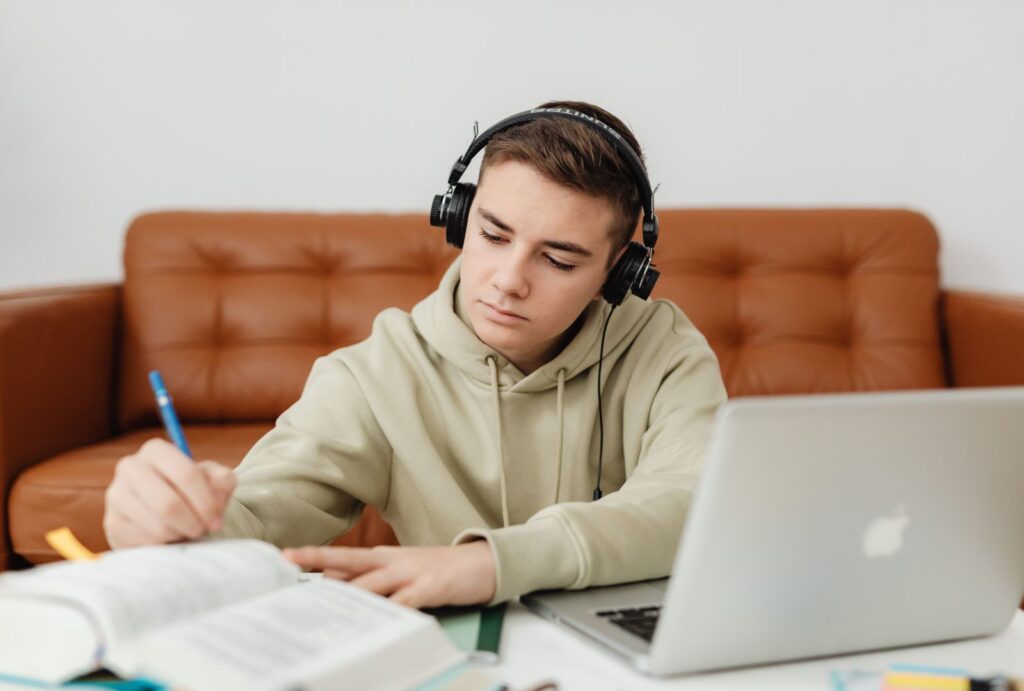 A focused young male student wearing headphones studies Hebrew diligently on his laptop, surrounded by open books, notebooks, and a blue pen, embodying the immersive online learning journey with Online Hebrew Teacher Anat Migdalor.