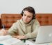 A focused young male student wearing headphones studies Hebrew diligently on his laptop, surrounded by open books, notebooks, and a blue pen, embodying the immersive online learning journey with Online Hebrew Teacher Anat Migdalor.