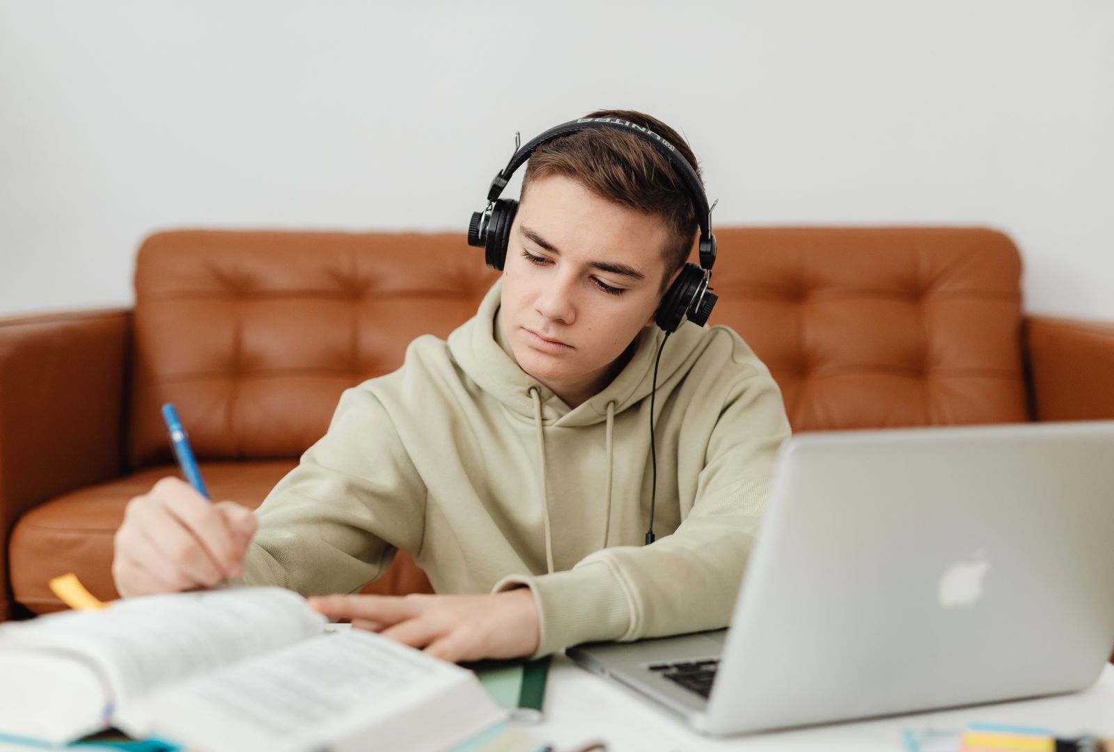 A focused young male student wearing headphones studies Hebrew diligently on his laptop, surrounded by open books, notebooks, and a blue pen, embodying the immersive online learning journey with Online Hebrew Teacher Anat Migdalor.