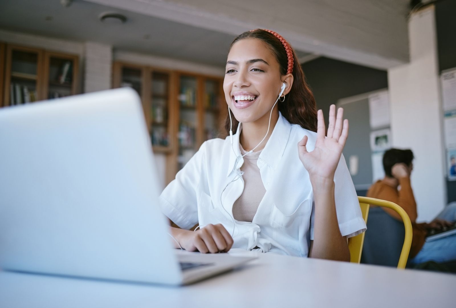 Smiling online Hebrew teacher with headphones waving hello during a virtual Zoom lesson on Hebrew language classes, laptop open on desk in cozy library setting.