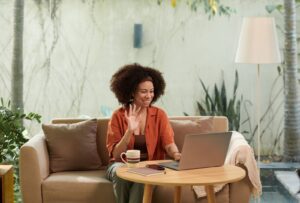 A smiling student learning Hebrew online via Zoom with her teacher Anat Migdalor, sitting on a cozy sofa with a laptop and coffee.