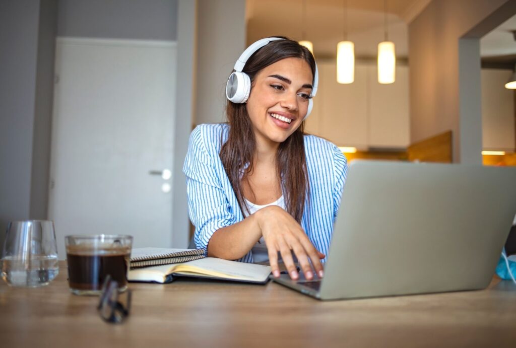 Woman wearing headphones studying Hebrew online at home with laptop and notebook on desk